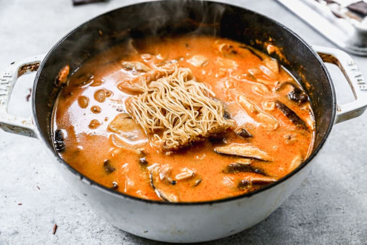 A pot of creamy, orange-red broth filled with mushrooms, garlic, and other ingredients, topped with a nest of uncooked noodles. The pot sits on a light gray surface, and steam is rising from the hot soup, indicating it is freshly cooked.