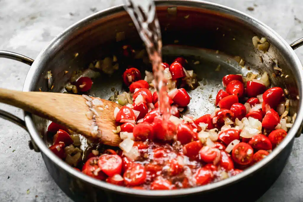 White wine being poured into burst cherry tomatoes and onions.